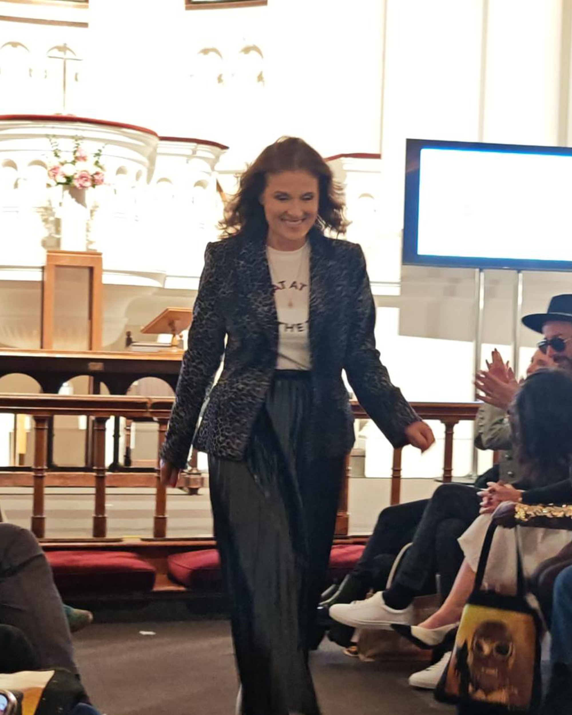 Woman in a suit walking in an indoor setting with people and a screen in the background