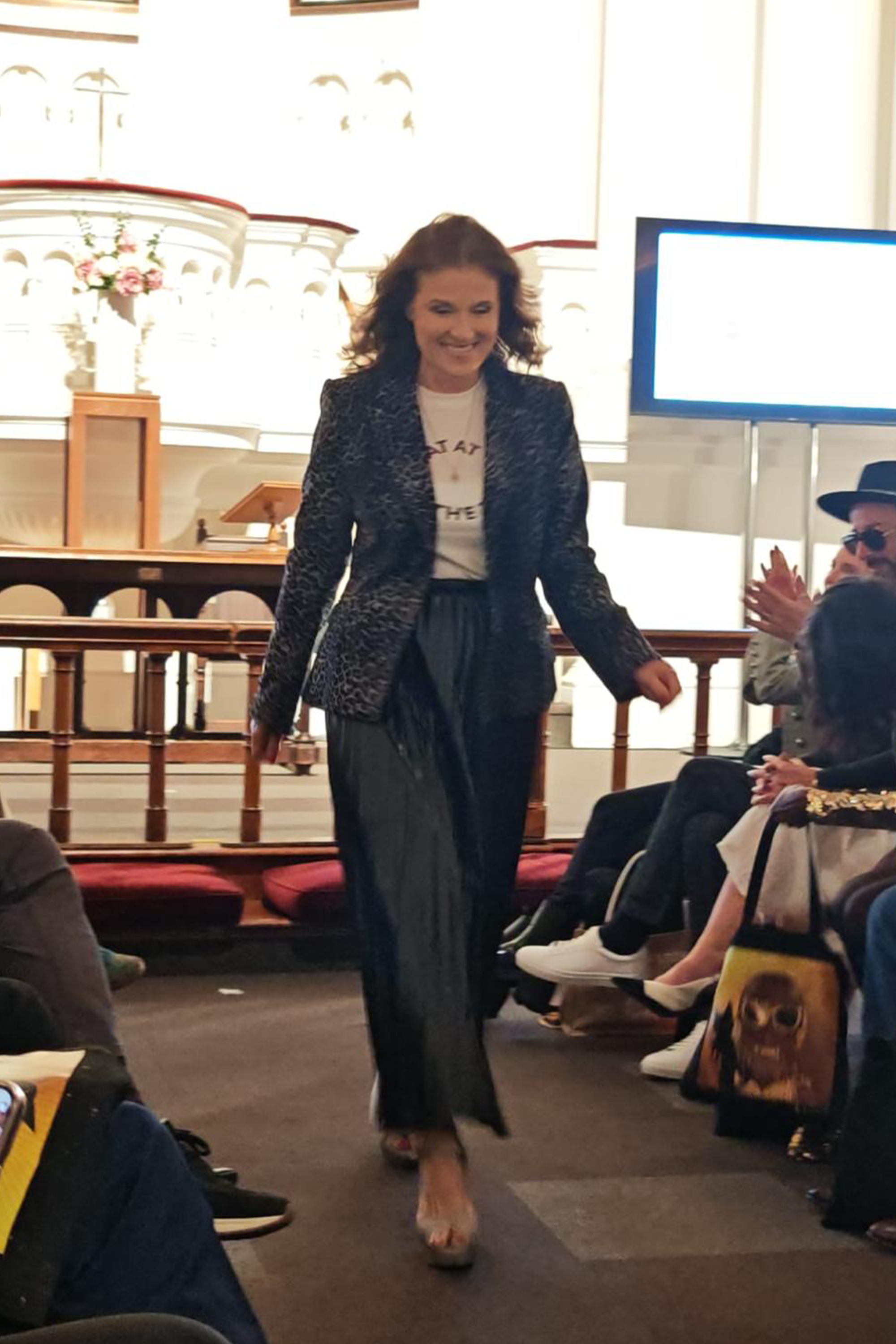 Woman in a suit walking in an indoor setting with people and a screen in the background